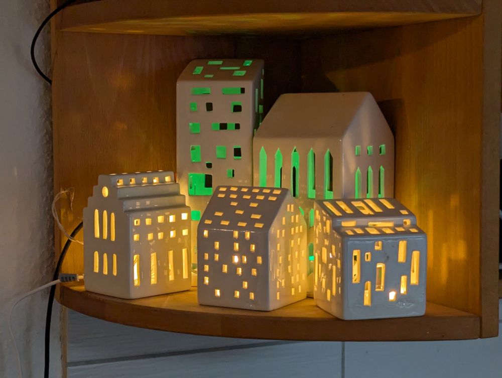5 Porzellanhäuschen in einem Eckregal sind beleuchtet. 3 niedrigere vorne warmgelb, zwei höhere dahinter grün aus den Öffnungen.