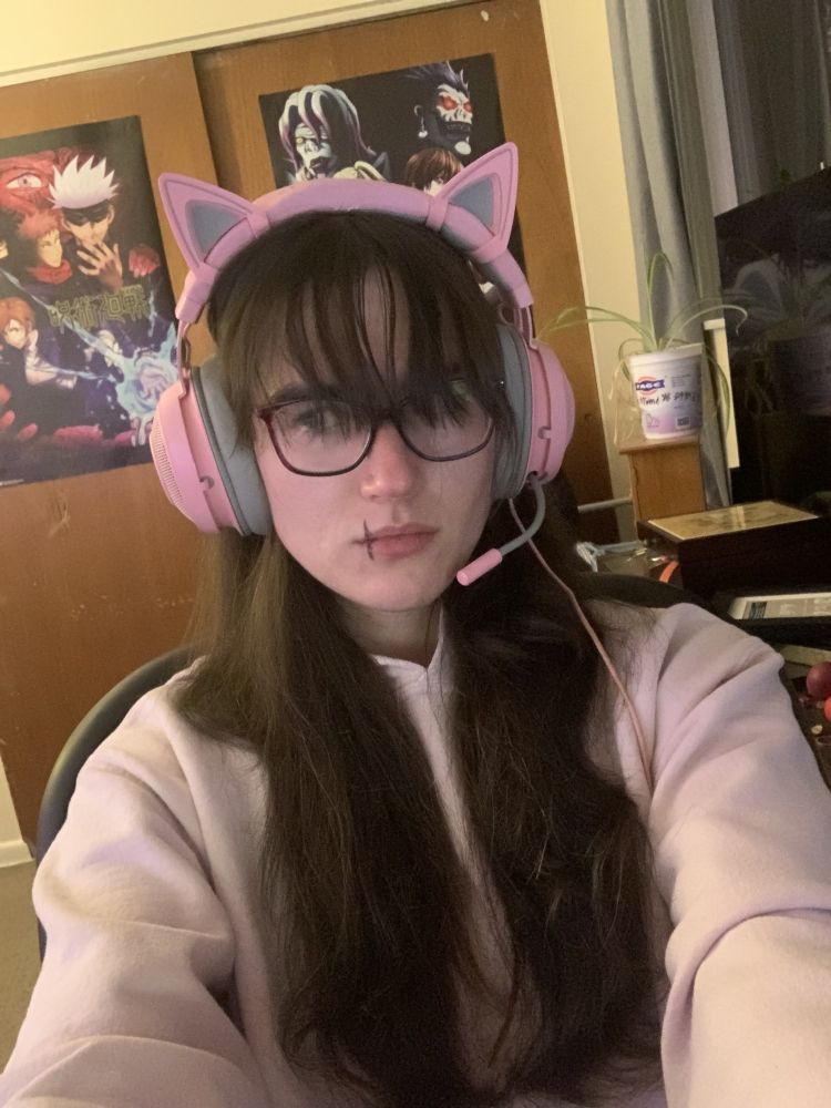 A white woman with long brown hair, a hoodie, and pink headphones. She has a makeup scar that looks like Toji from JJK.