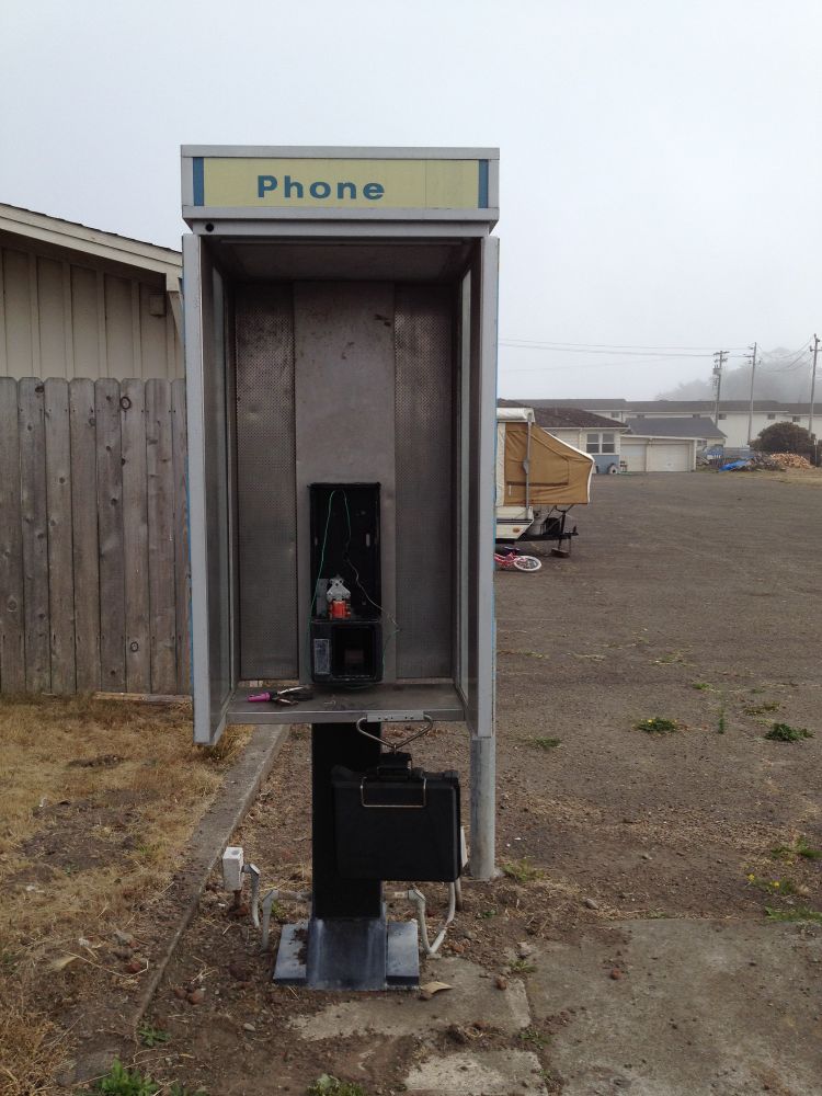 Eine verlassene Telefonzelle ohne Telefonapparat steht auf einem unbefestigten, teilweise betonierten Boden neben einem alten Holzzaun. Die obere Leiste der Zelle trägt die verblasste Aufschrift „Phone“. Im Inneren der Zelle sind nur noch Kabel, ein leerer Metallrahmen und einige übrig gebliebene Bauteile zu sehen. Auf der Ablagefläche liegt ein rosafarbener Gegenstand, möglicherweise ein Feuerzeug. Unten am Sockel hängt ein schwarzer Kasten mit einem offenen Metallbügel, aus dem weiße Kabel herausführen. Im Hintergrund befinden sich ein Wohnwagen, ein Kinderfahrrad, einfache Gebäude und ein trüber Himmel, der eine melancholische, fast verlassene Atmosphäre unterstreicht.
