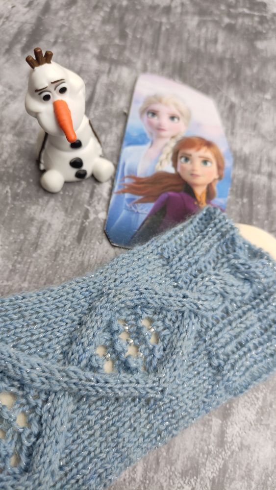 Cabled sock a bit in the look of a window with beads added within that frame. Glittery  light blue yarn in the front, mainly shaft show on a sock blocker. An Elsa and Anna game card as well as an Olaf tonie in the background.