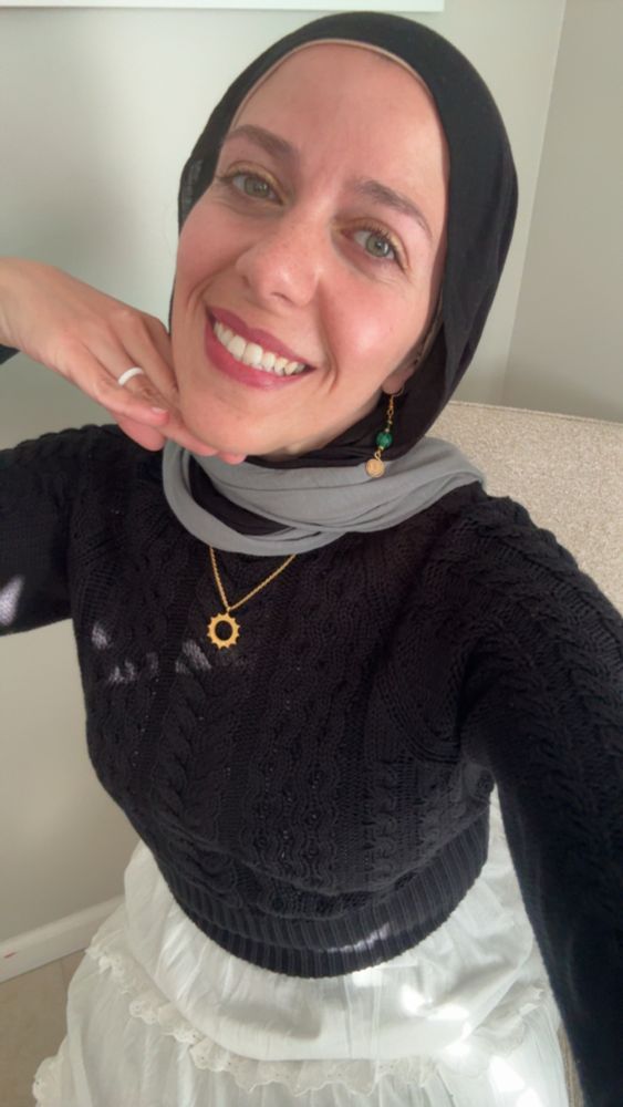 Aya smiling big wearing a black and gray hijab sitting on a cream chair and wearing a black sweater and white skirt. She has gold earrings and necklace 