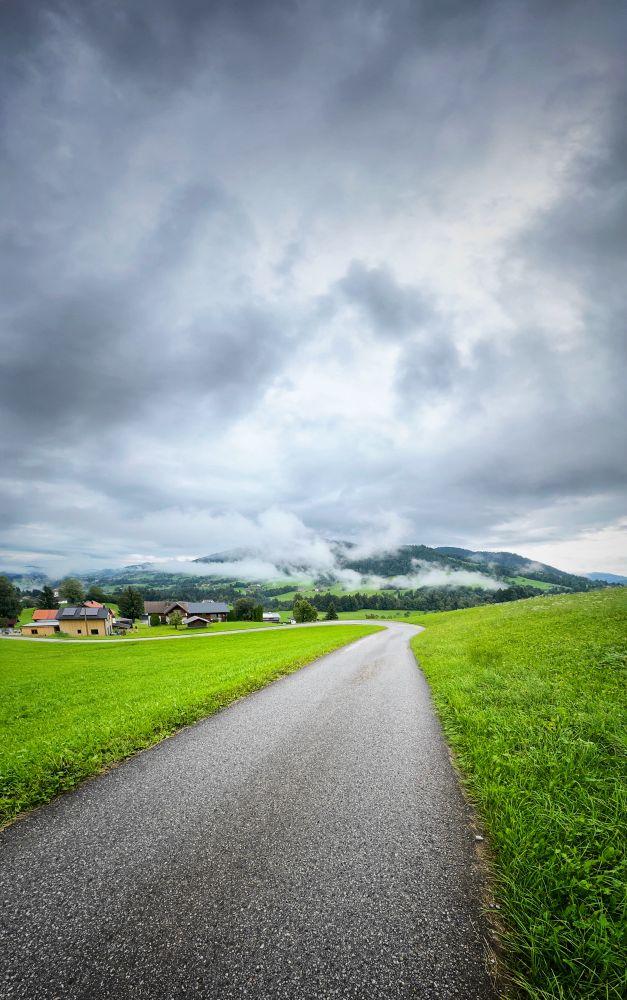 A narrow paved road winds through a lush green meadow in a countryside setting. On the left side of the image are a few scattered houses with red and dark rooftops. In the distance, low-lying clouds and mist hang over rolling hills covered in green forest. The sky above is overcast with dramatic gray clouds, creating a moody, atmospheric scene