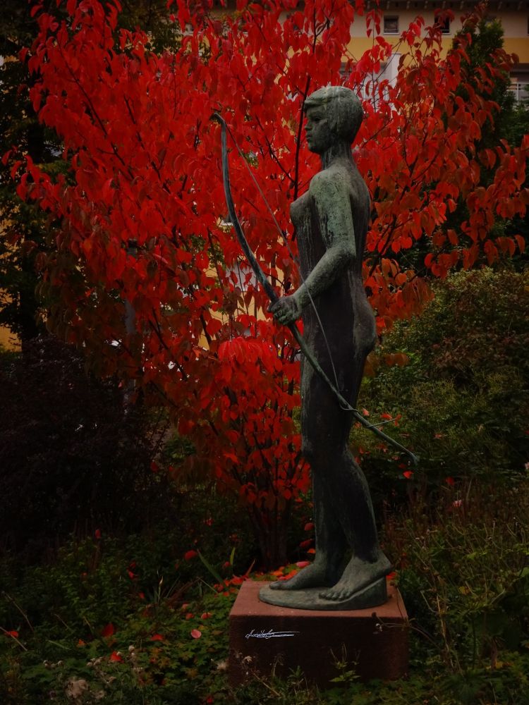Die mit Grünspan bedeckte Bronzestatue einer stehenden nackten Frau mit kurzen Haaren. Die Figur ist von der Seite fotografiert. In der linken Hand hält sie einen Bogen. Den rechten Arm sieht man nicht. Hinter der Statue wächst herbstlich strahlend rot ein Busch in die Höhe. Dahinter grüne Hecke, darunter grüne und braune Wiese auf der einige rote Blätter vom Busch liegen. Die Statue steht auf einem rechteckigen rostbraunen Sockel.