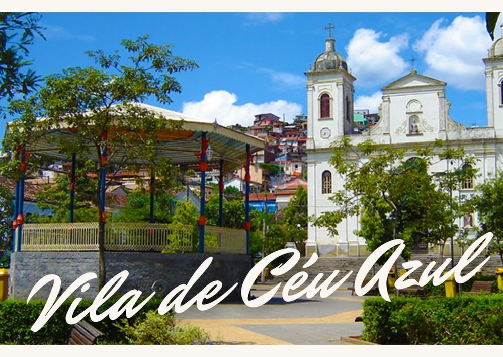 Frente de um cartão postal, com uma foto de uma pracinha de cidade pequena, com um coreto e uma igreja. Em letra cursiva está escrito "Vila de Céu Azul"