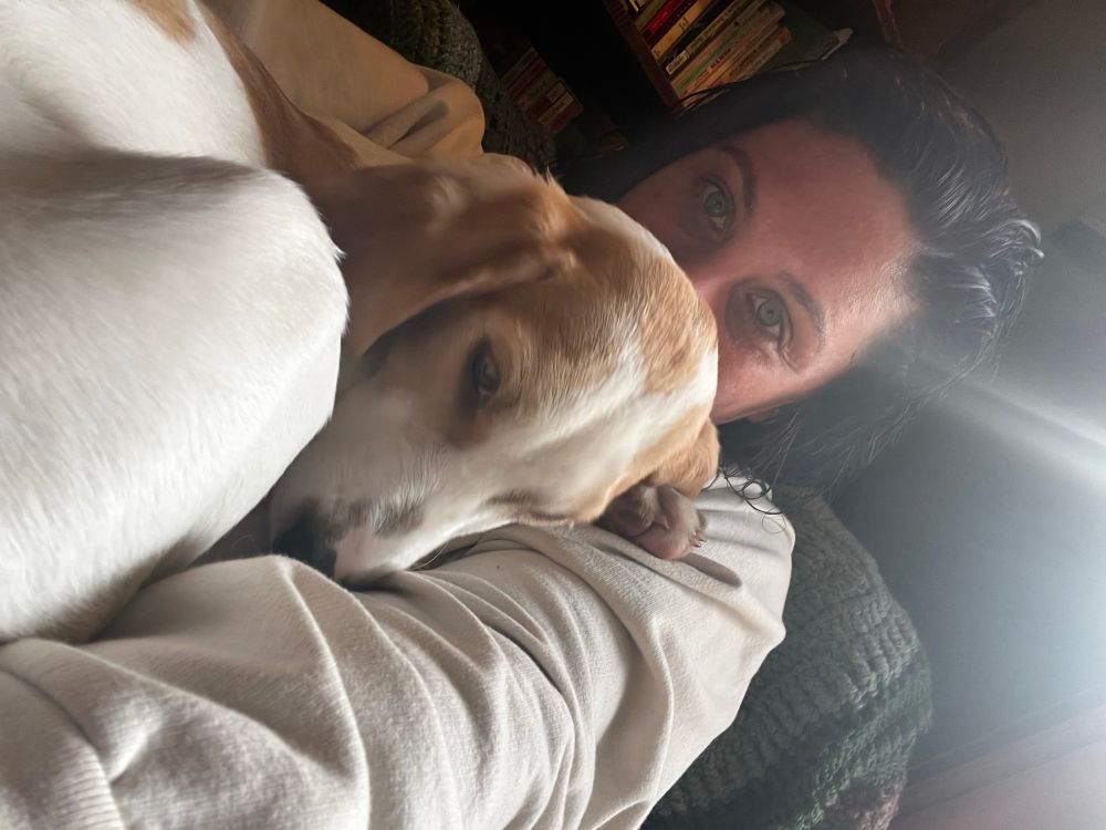 A snuggly lemon foxhound puppy in the foreground is trying to sleep while a dark haired person hugs her and kisses the back of her head.