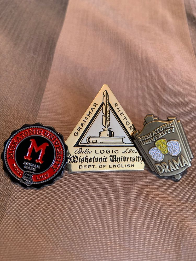 Three lapel pins, one for Maskayonic University alumni, one for the Miskatonic U Department of English, and one for the Miskatonic University Department of Drama.