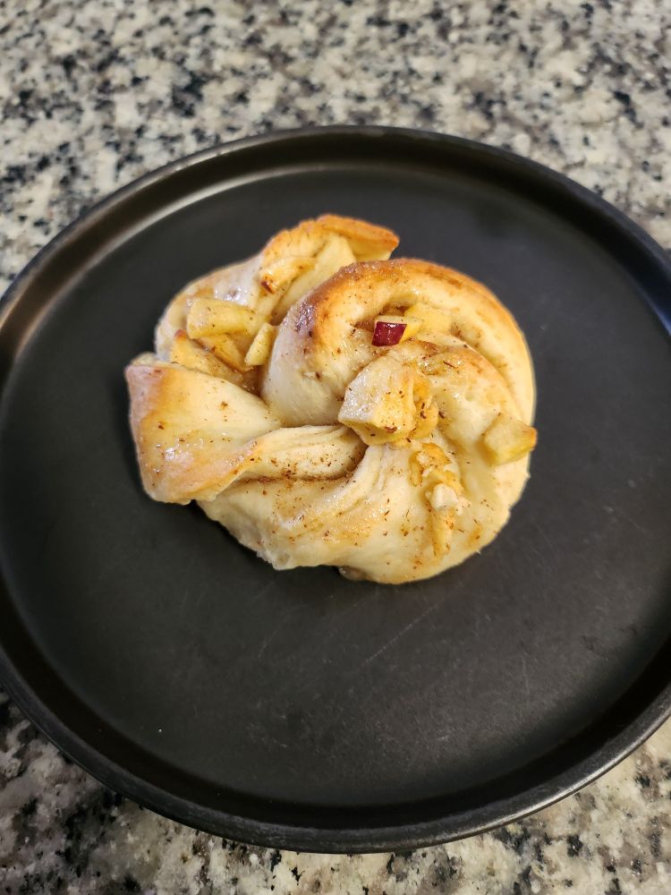 A bread knot with apple and cinnamon baked to golden brown resting on a black plate.
