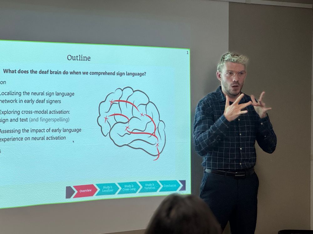 A white male presenting person with short blond hair, wearing black pants and dark check shirt signing ASL and standing in front of a projection screen showing a drawing of a brain with black text on a white screen.