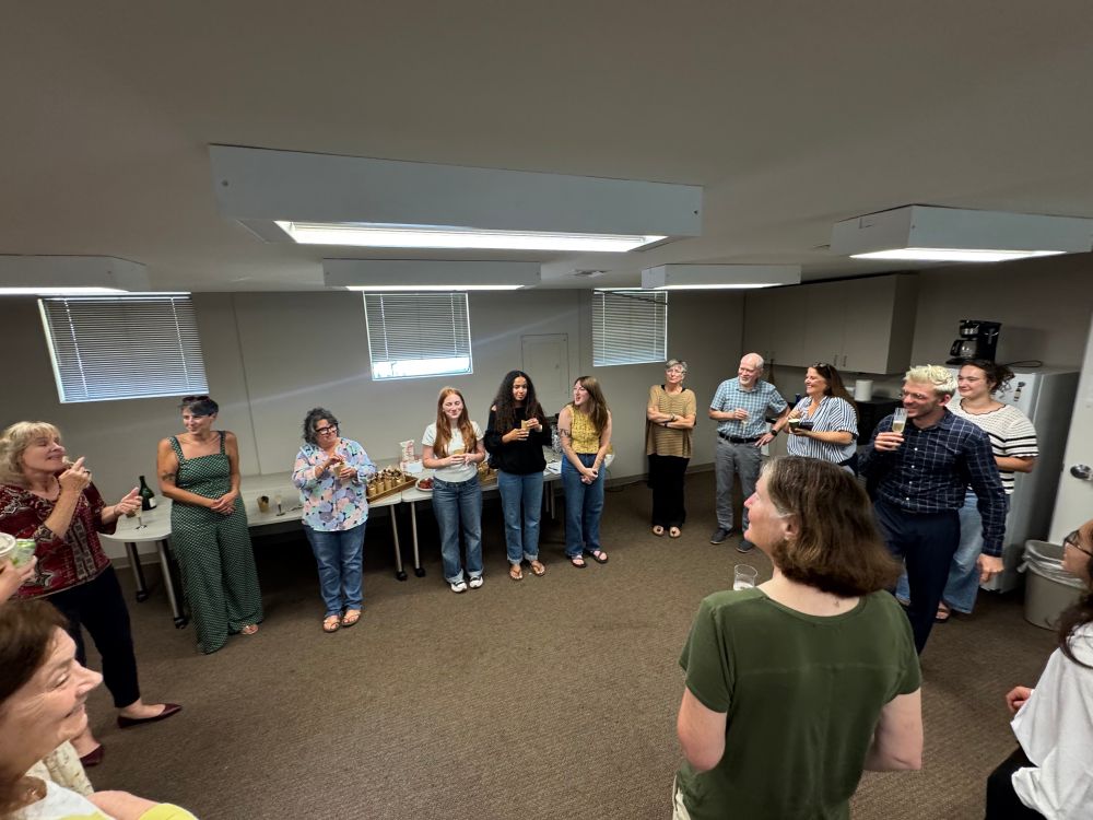 A group of people standing in a circle im a conference room with the window blinds drawn, one female is signing ASL to the rest of the group