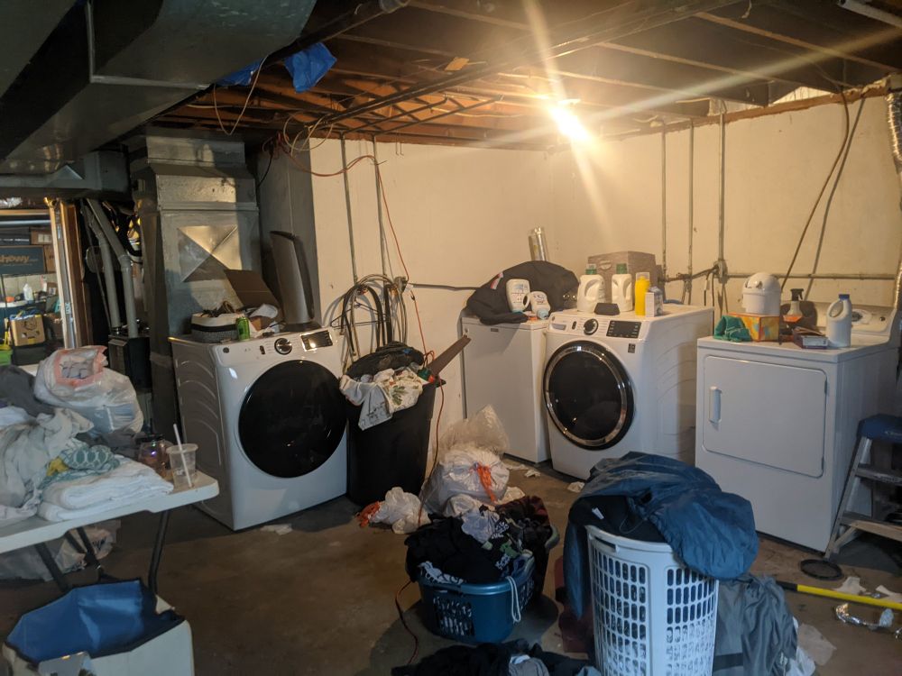 Unfinished basement room with two sets of washers/dryers, unfinished walls and a bunch of laundry.