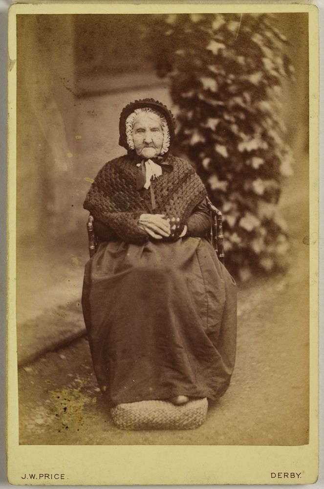 Anne Banton sitting in a wood chair, wearing a black dress, shawl and bonnet, outside Calke Abbey. Her feet rest on a knitted cushion. There is a column of ivy behind her left shoulder. The cream surround to the photograph states that it was taken by J. W. Price of Derby. 