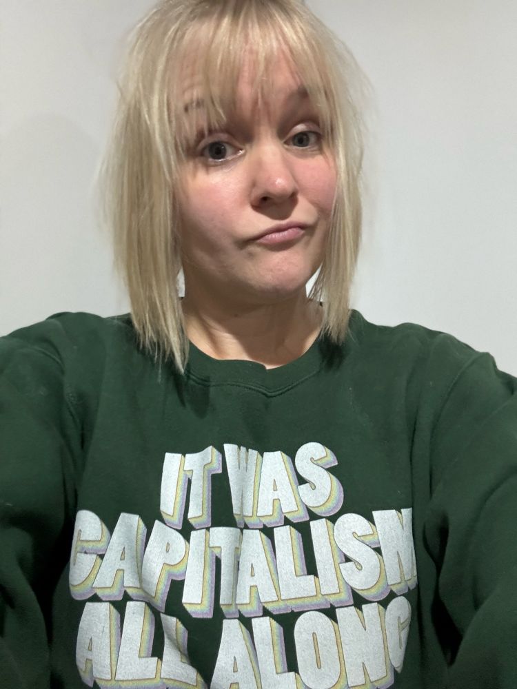 Selfie of a blonde, white woman making a sarcastic face while wearing a green sweatshirt that says “it was capitalism all along.”