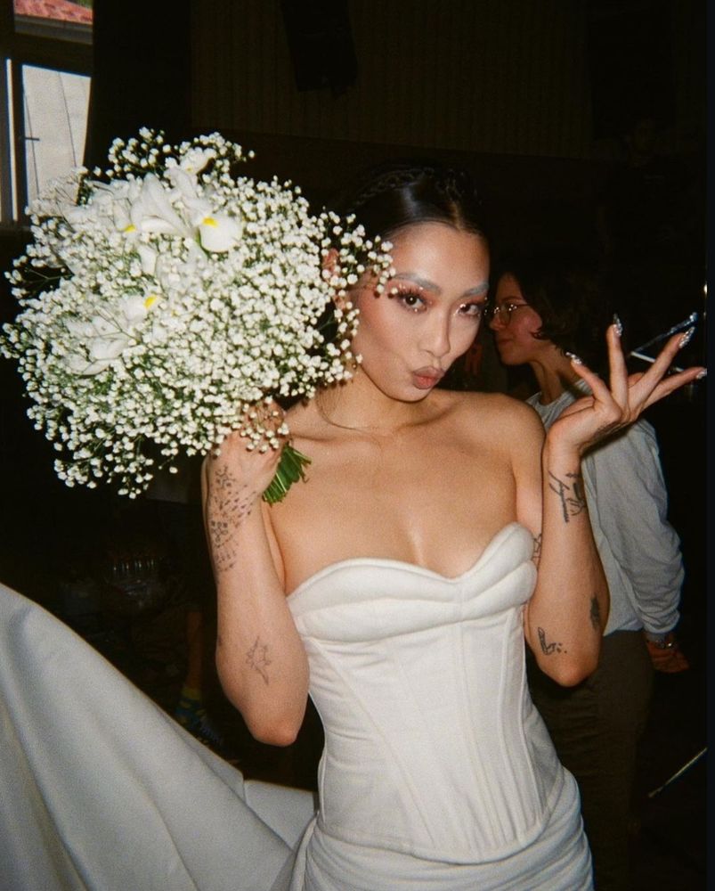 A photo of musician Rina Sawayama behind the scenes of filming the music video for her song "This Hell". She is wearing a white wedding dress and holding a white flower bouquet.
