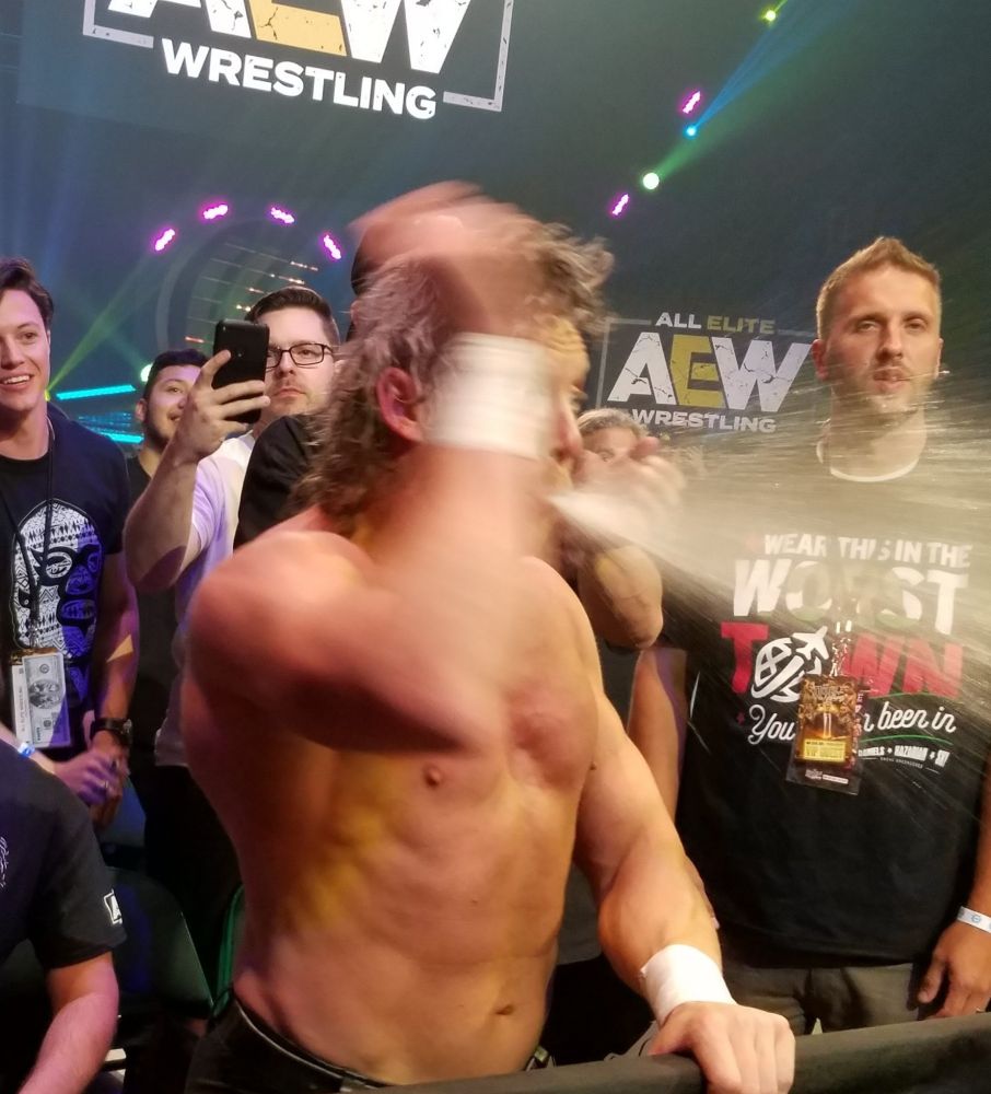 Kenny Omega, among fans at ringside, spits a large spray of water at an off-camera Chris Jericho.