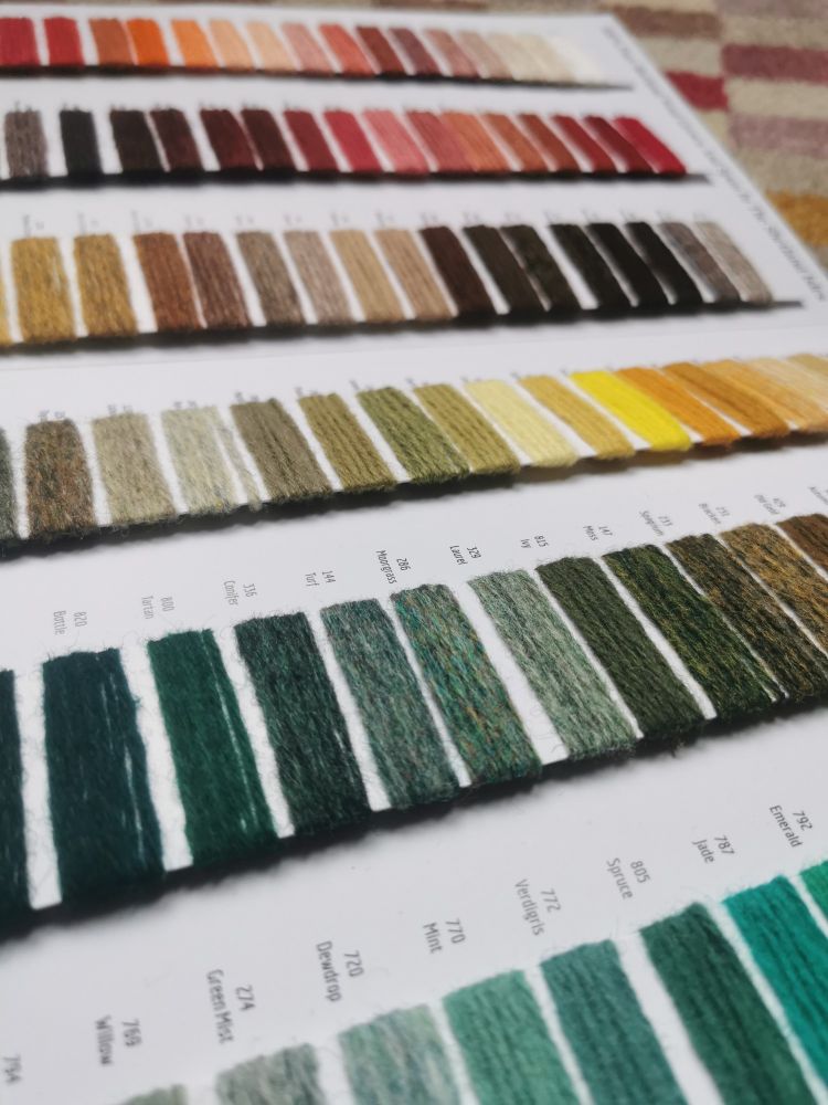 An opened out shade card of Shetland wool shows many wrapped yarn swatches in greens, yellows and pinks on white card strips. 