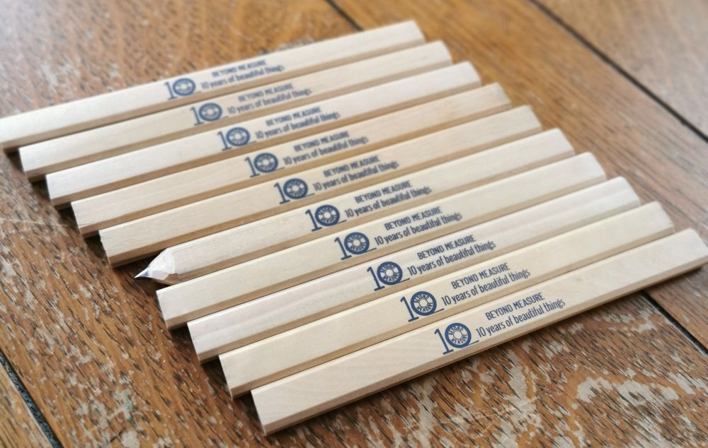 A photo of 10 natural wood carpenter pencils lying in a row on an old wooden table.
Each one is stamped with 'Beyond Measure 10 years of Beautiful Things'
One is sharpened, the others are blunt