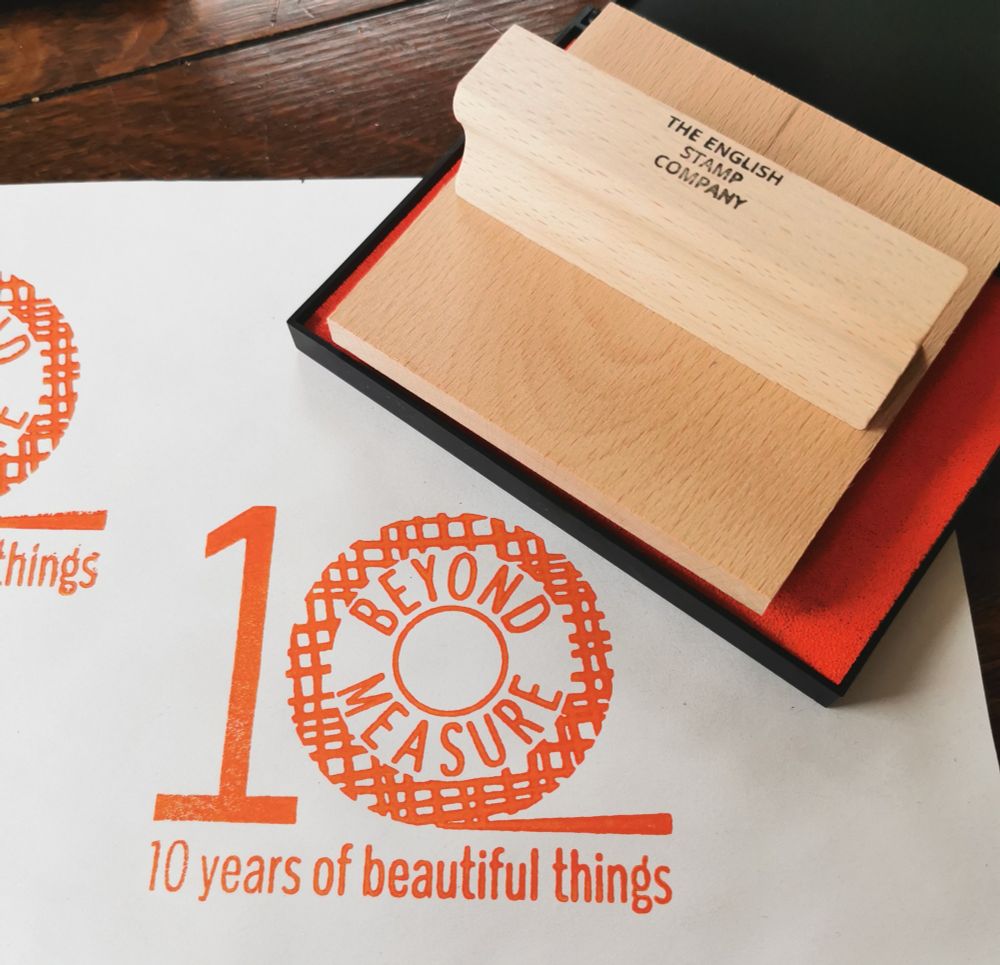 A large stamp of the Beyond Measure 10 year anniversary logo is printed in orange on a piece of paper.
An orange ink stamp is in the background.
Text beneath the logo reads 10 years of beautiful things. 