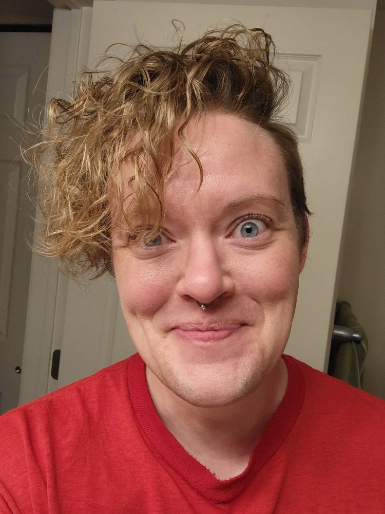 a head and shoulders image of a white non binary person with a goofy barely held in grin and blue eyes crinkled in a smile looking at the camera. They are wearing a red t-shirt and a mess of chaotic blonde curls flops over the head and across one eye.