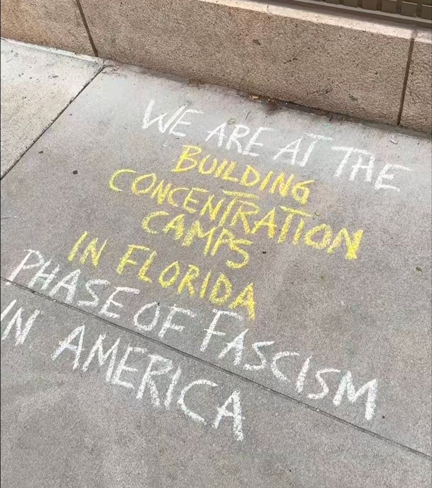 Chalk writing on a sidewalk that reads: we are at the building concentration camps in Florida phase of fascism in America.
