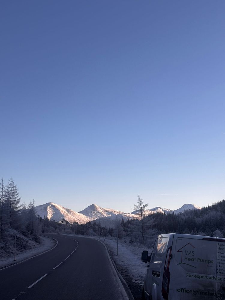 A scenic winter road winds through a snowy landscape with tall trees dusted in white, leading towards a backdrop of the snow-capped Cairngorm mountains glowing in soft morning light. The sky is a clear, vivid blue. On the side of the road, an IMS Heat Pumps van is parked, with the company logo and contact details visible. 