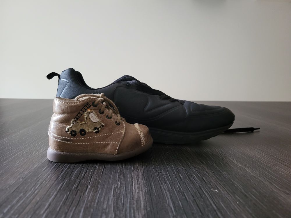 Une chaussure de bébé marron avec 1 camion de pompiers marron aussi est posée sur une table. Derrière, alignée au niveau du talon 1 basket noire 2 fois plus grande...