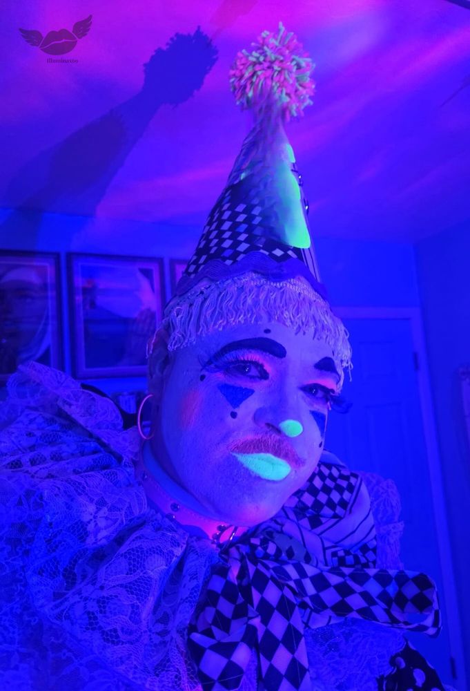 Illuminax sharing a selfie dressed up as a clown wearing a lace, checkered neon outfit with a cone shaped hat and neon glowing clown makeup in room with pink-purple lighting.