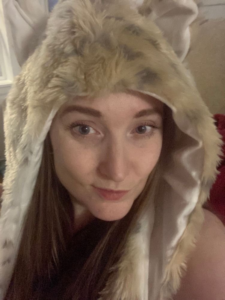 A pale woman with long blonde hair in a fuzzy animal hood with ears. My cheekbones are, to be quite honest, fabulous. You could ski jump off of them. You could slit a throat on their sharpness. 