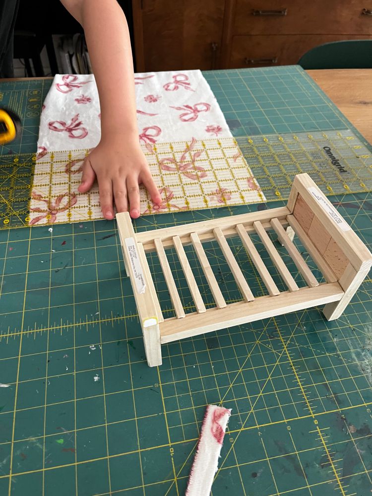 A 7”x4”x3” doll bed made of wood. Daughter is cutting a piece of fabric in the background as a blanket. 