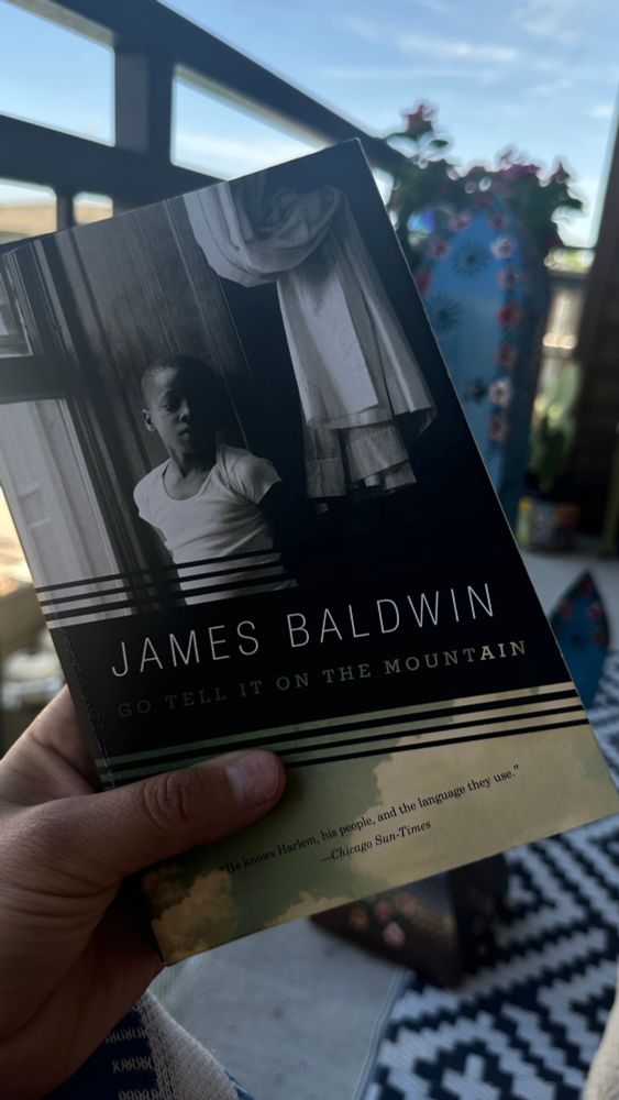 Kolbe holding a copy of James Baldwin’s Go Tell It On The Mountain. The background is a blurred depiction of some flowers and balcony decorations 