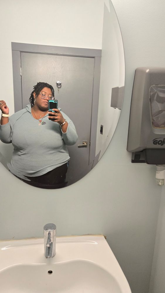 A bathroom mirror selfie of me (brown transmasc) wearing a pair of black leggings and a blue long sleeve shirt with a hood. 