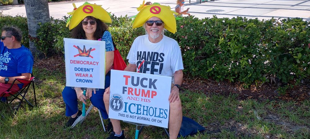Couple sitting in the shade wearing chicken taco hats with "Tuck Frump" and "No Kings" signs.