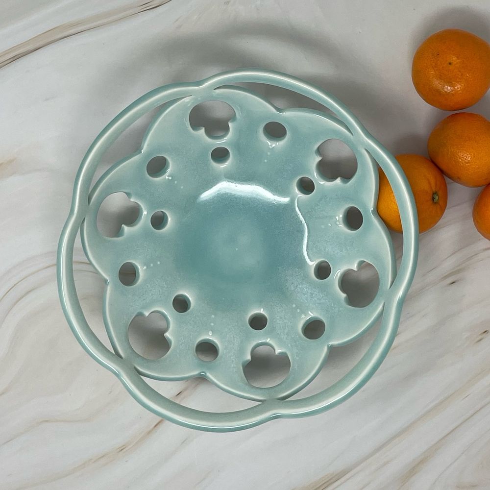 Soft blue celadon bowl with pierced and cutout negative space design, shown on marble countertop with orange mandarins