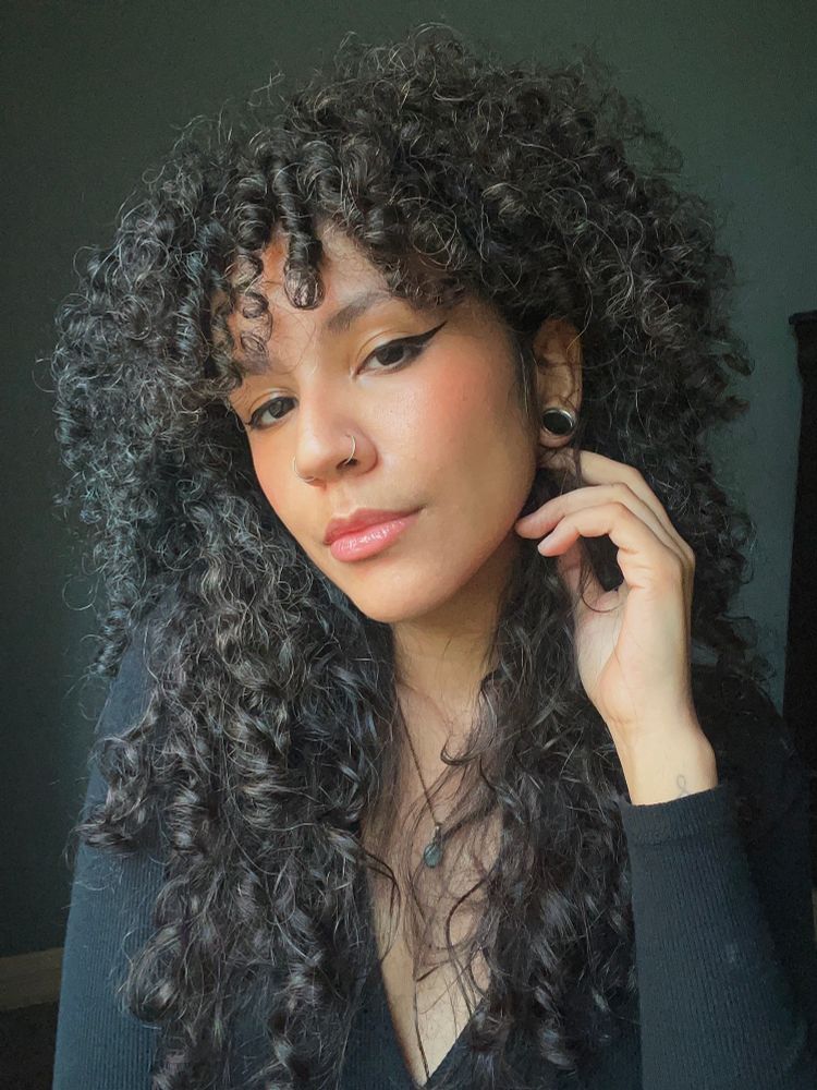 a Sopa selfie where she’s looking into the camera at a 3/4 profile. They have long black curly hair, light olive toned skin, stretched ears, and double nostril piercings with hoops. She’s wearing a long sleeved black shirt and larimar crystal tear drop necklace. Her expression is serious.