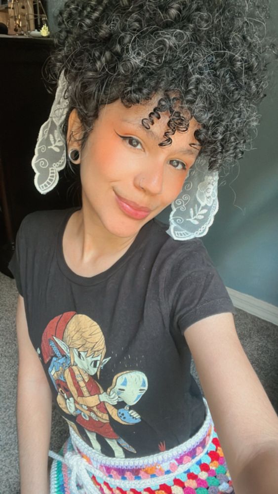smiling selfie of Sopa. She has black curly hair pulled into a ponytail, has a lace ribbon in their hair, and is wearing a Legend of Zelda and Spirited Away crossover t-shirt.