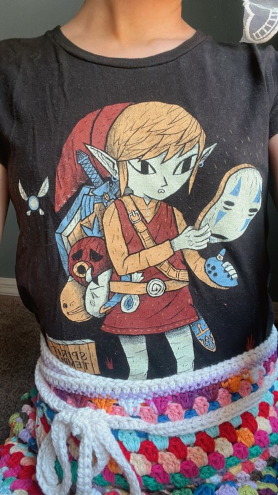 a close up shot of Sopa’s Legend of Zelda and Spirited Away crossover t-shirt. A stylistic 2D Link is holding an ocarina and a mask of the character No Face. Sopa is also wearing a colorful crochet wrap skirt she made herself.