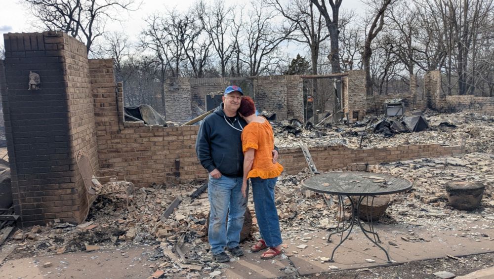 Andrine and her husband kevin stand in front of what is left of their burned house. There's mostly piles of bricks and one metal table.