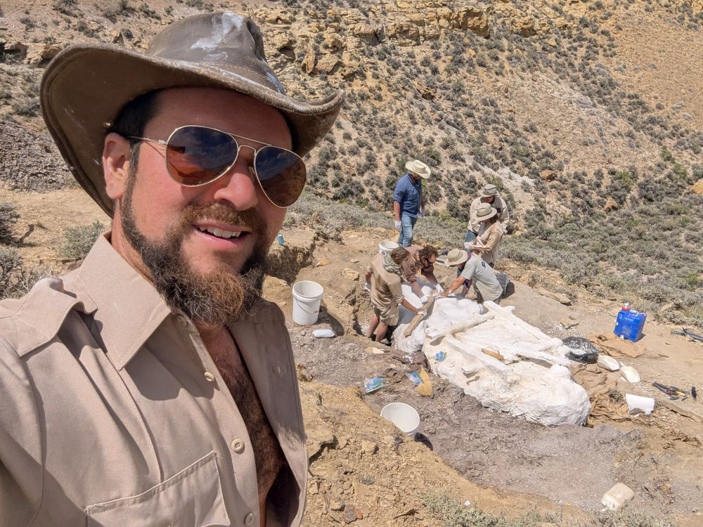 Selfie photo of me in front of the hadrosaur quarry.  Behind stand six members of our crew around two huge plaster field jackets 