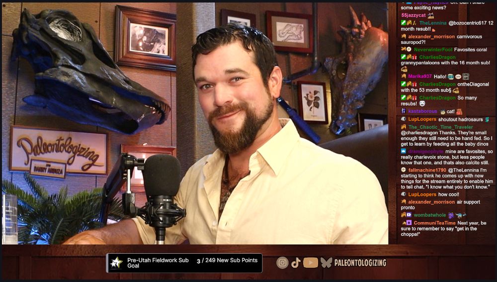 Twitch screenshot of me in my office.  Behind me are life-sized 3D prints of a Diplodocus and Pachycephalosaurus skull on the wall, as well as framed prints of Barnum Brown, hadrosaur elements, etc.