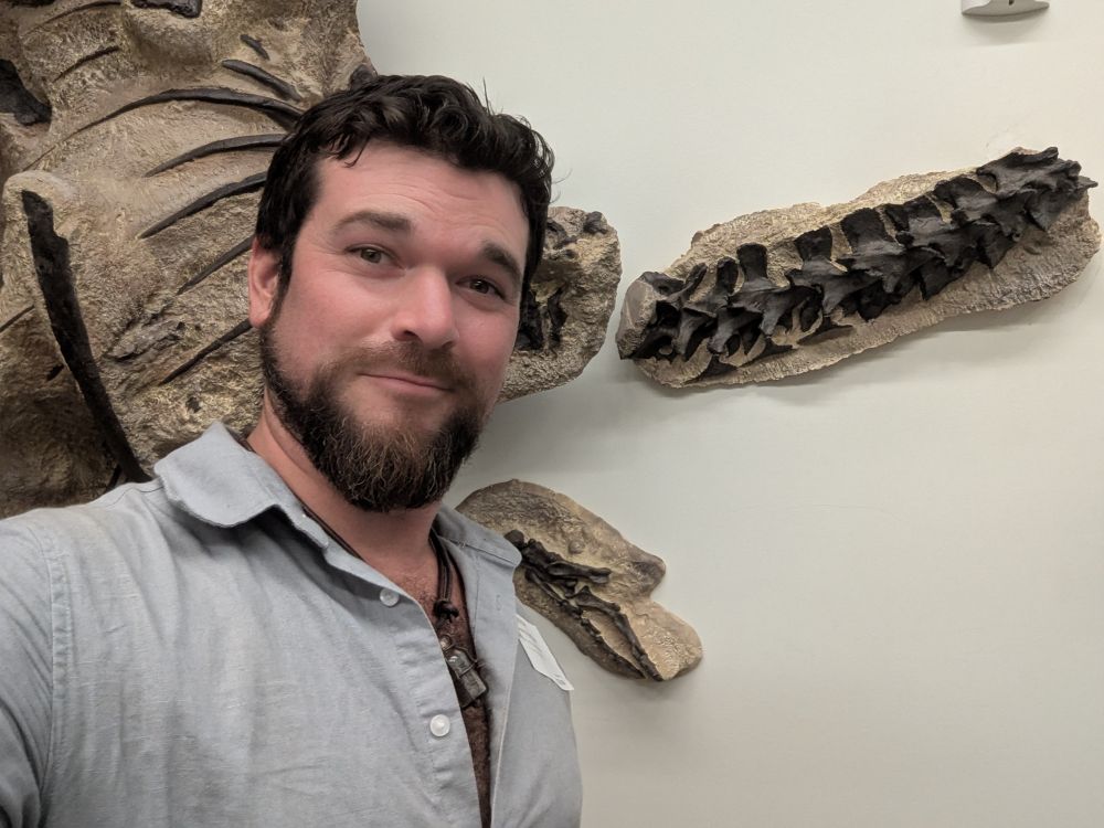 Selfie photo of me in front of the Allosaurus skeleton, mounted to the wall inside NHMU's vertebrate fossil collections facility