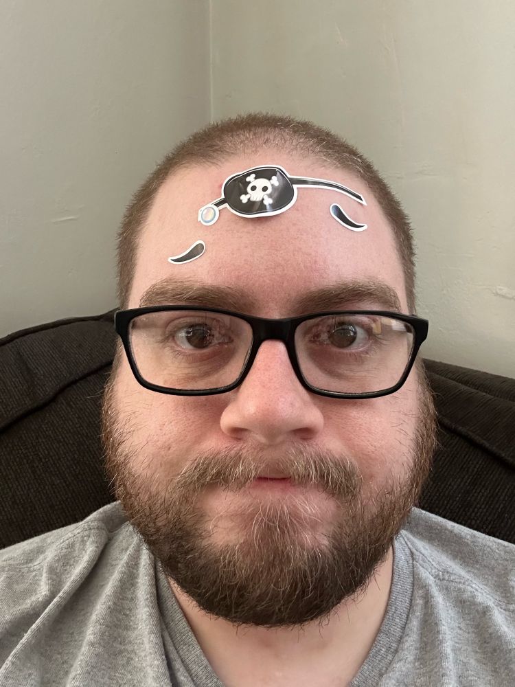 A mid-30s man with assorted pirate themed stickers applied by his sons to his forehead with an “unamused” look on his face. 
