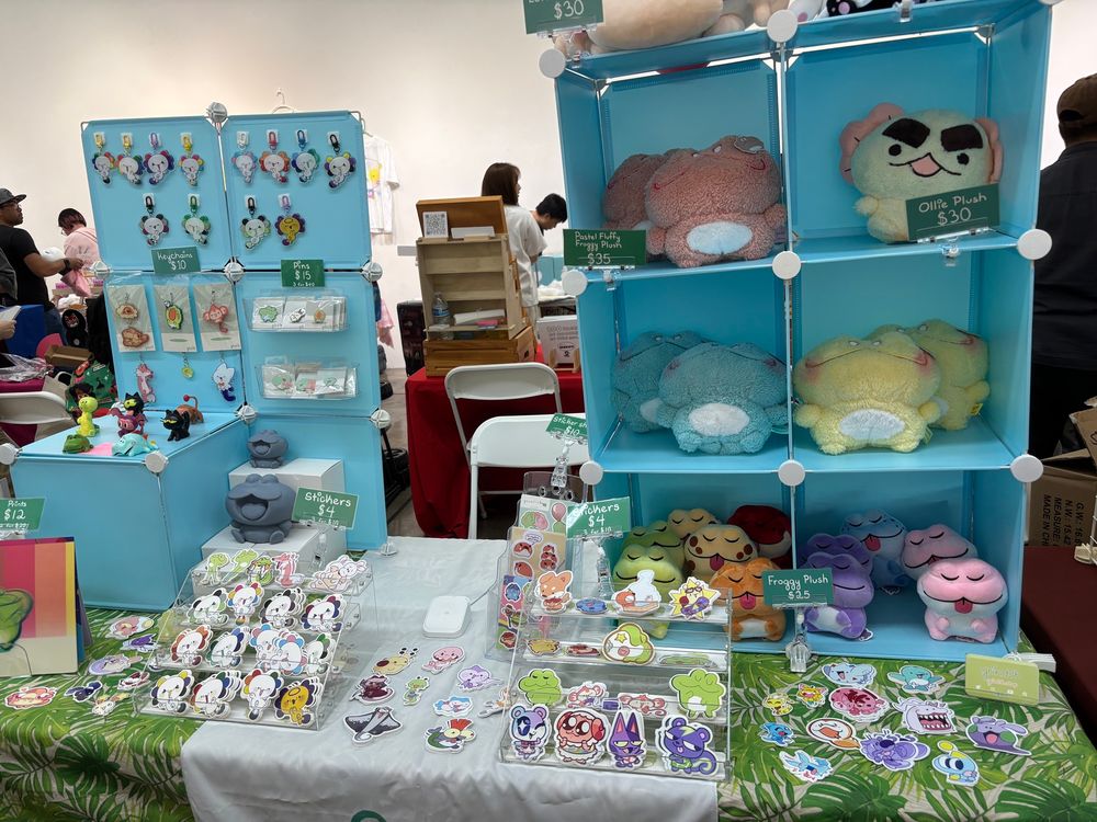 A light blue convention booth featuring colorful frogs, some axolotls, ghost cats, tons and tons of other various critters.