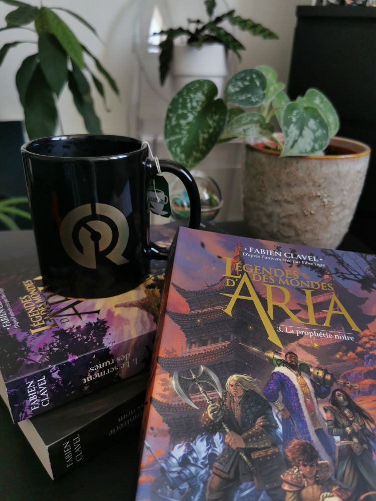 Une tasse noire avec le logo de l'émission Game of Roles est posée sur deux livres dont les couvertures ne sont pas visibles. Au premier plan, le livre « Légendes des mondes d'Aria ; La prophétie noire ». La couverture représente 4 personnages dont un tient une hache double et un autre vêtu d'un grand manteau violet tient une seringue géante remplie d'un liquide vert. Derrière les personnages se dessine un décors dans des tons rosés et bleus.
En arrière plan de la photo on peut distinguer plusieurs plantes vertes posées sur des meubles.