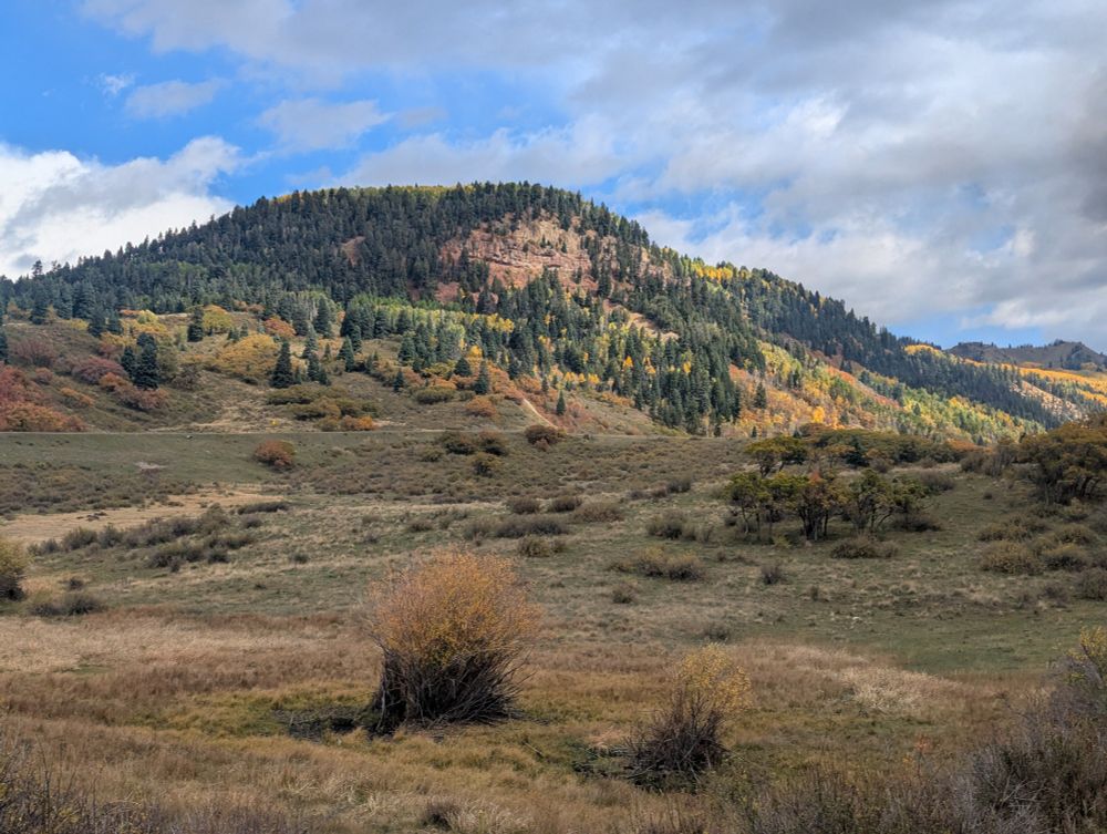 A partly cloudy sky highlights a rock face covered in conifer and bright yellow aspen.