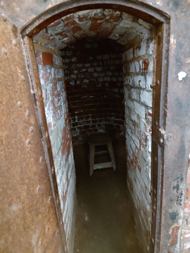 Photo of a solitary confinement cell, referred to as "the hole". The room is tiny - the entrance is barely wide enough to fit someone's shoulders, and low enough that most adults would have to stoop to enter. The walls are made from crumbling brick. A little further in is a tiny round room, maybe four feet wide at most, with a wooden stool in the middle. There is no light inside. The door is made from heavy iron, now rusted, and would probably leave someone in near total darkness once closed.