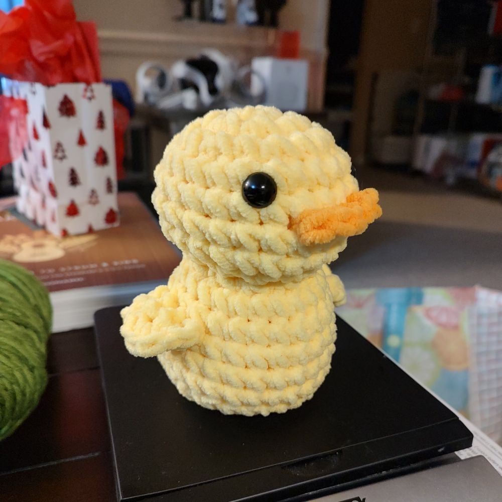 Photo of a crocheted duck plushie. It's made with a fuzzy chenille yarn. The body is all yellow with an orange bill and black doll eyes.