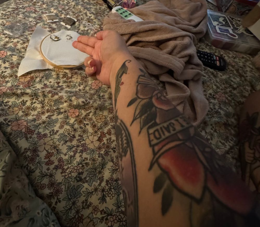a tattooed arm with hand formed into finger guns pointing at an unfinished cross stitch in an embroidery hoop. the hoop is on a bed with floral sheets. 
