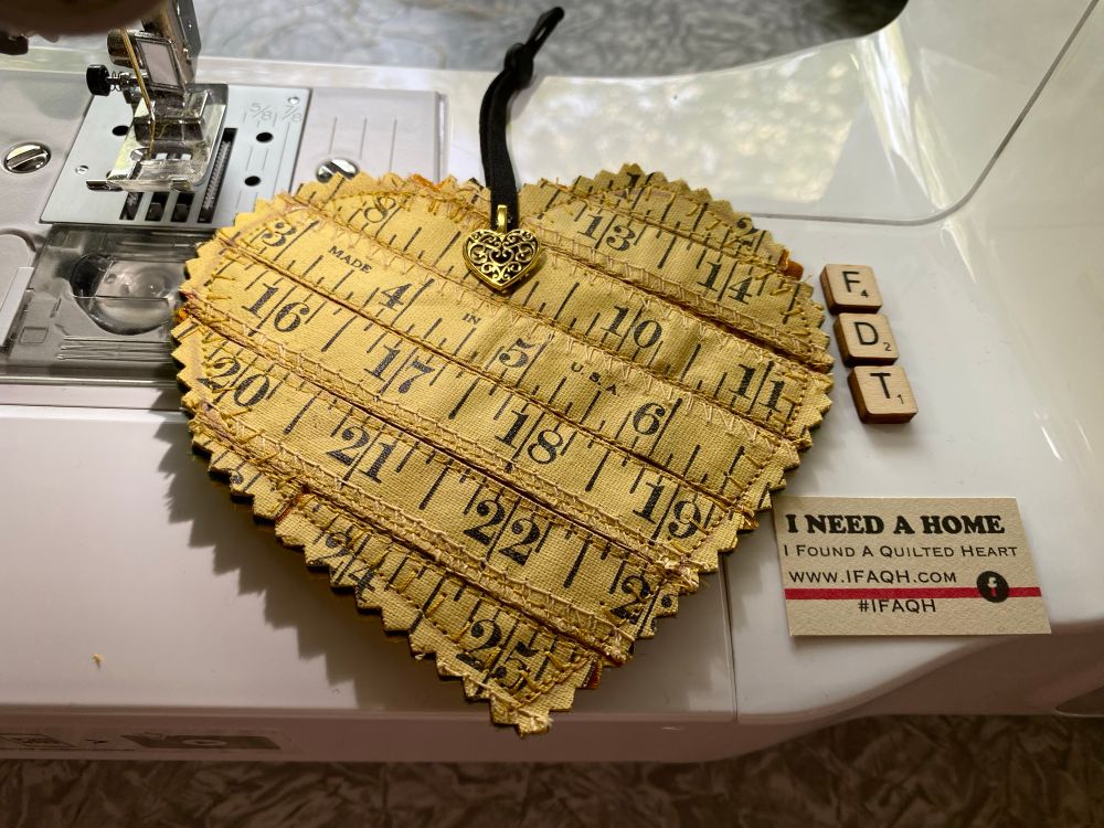 The image shows a heart-shaped fabric ornament placed on the plate of a sewing machine. The ornament is made from strips of a yellow measuring tape, arranged horizontally and stitched together. The fabric has black measurement markings (in inches), and pinked edges (zigzag cut) for a decorative touch. It states that it was made in the USA. At the top of the heart is a black ribbon loop with a small gold heart charm attached, which has a filigree design.

The heart is positioned under the presser foot of the sewing machine, suggesting it may have been recently sewn. The overall aesthetic blends sewing-themed materials with a handmade, vintage charm.