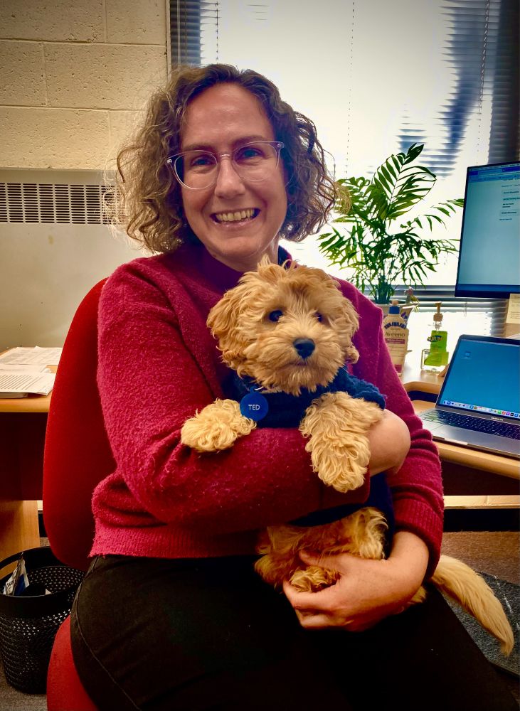 Kate Bagnall pictured sitting in her university office holding a small and very cute dog whose name-tag says ‘Ted’.