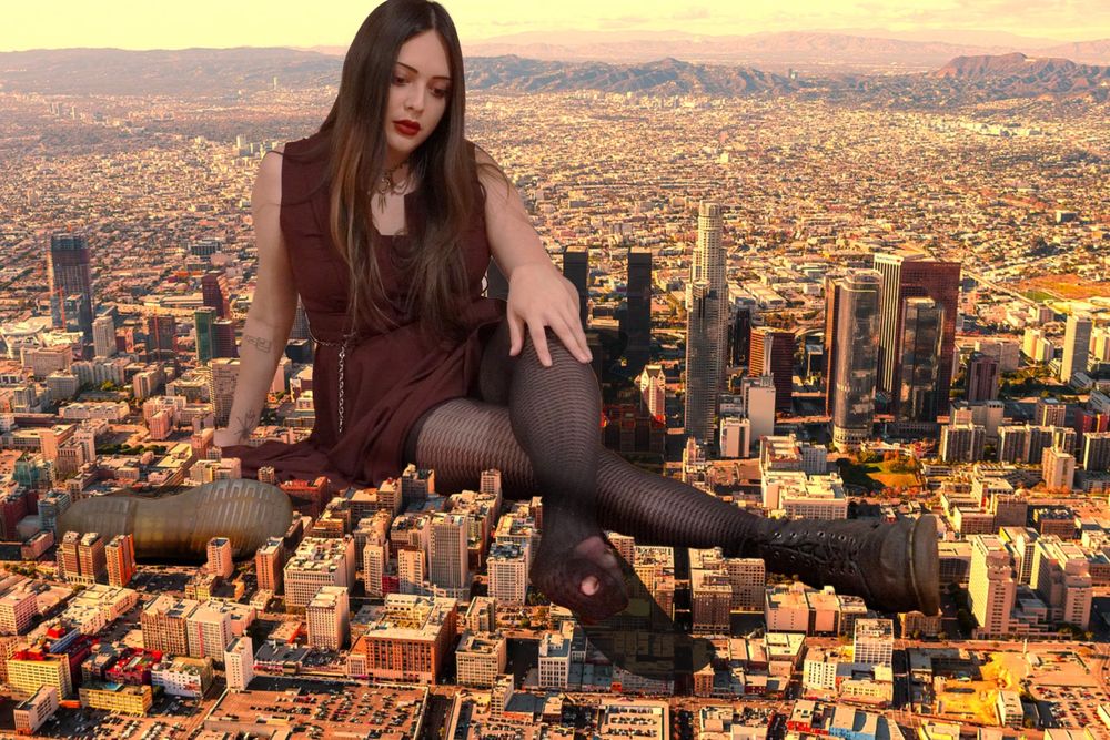 me, a giantess with long straight brown hair, brown eyes, and dark berry red lipstick, wearing a short burgundy dress with a chain belt, sheer tights, and one Doc Marten boot on my right foot, sitting in the middle of a city with my legs stretched out, the highest skyscraper not even reaching my shoulder. my left foot has no shoe and there is a small hole in my tights near my toes, it covers over a street and casts a shadow across the city block which i could easily level in a single step. my left boot sets next to me on its side, sole to the camera, dwarfing the dozen-story buildings. the city is mine.