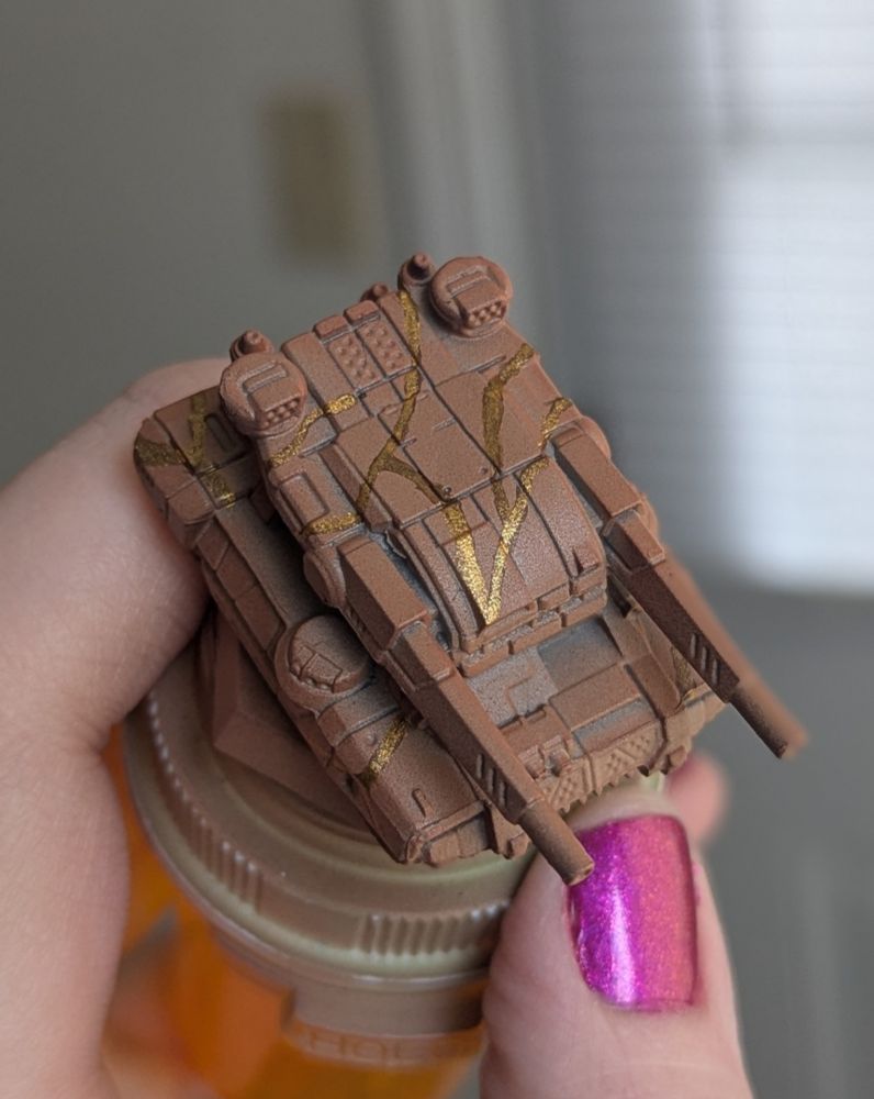 A miniature twin barreled tank with a large, flat turret painted in terracotta brown with a gold kintsugi pattern.